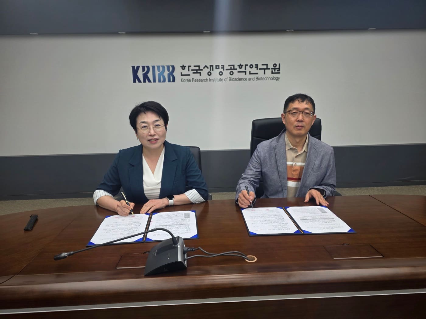 Two people seated at a conference table signing agreement folders, in front of a wall bearing the KRIBB logo and its name in Korean and English.